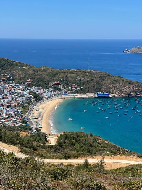 A fishing village on a curved bay, temples on the hill and blue boats on turquoise water, seen from a hillside path — Original