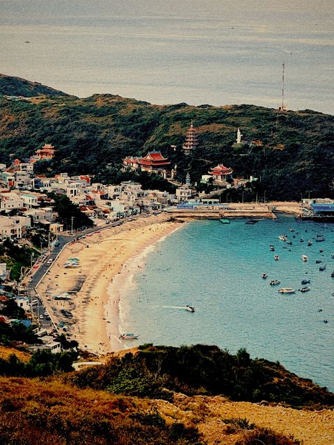 Curved sandy bay below a green hill with red-roofed pagodas, boats on turquoise water — shot on ELEV ARCADE