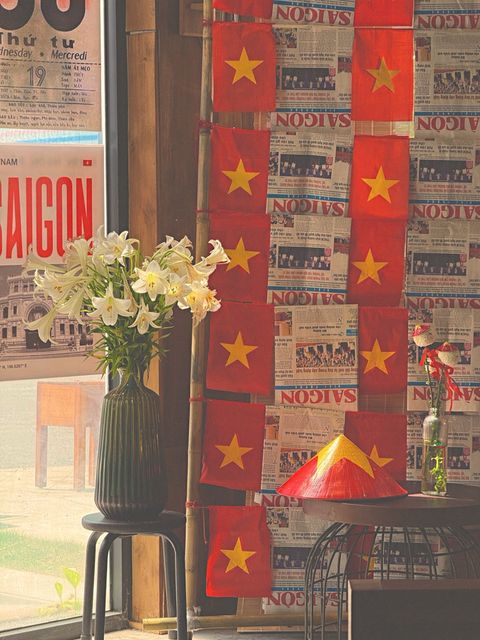 A café wall of red flags and old newspaper pages, white lilies in a vase by the window — shot on ELEV 01