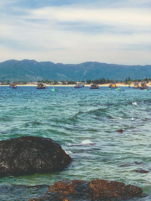 Fishing boats anchored off a beach, dark rocks in front and mountains beyond — shot on ELEV 04