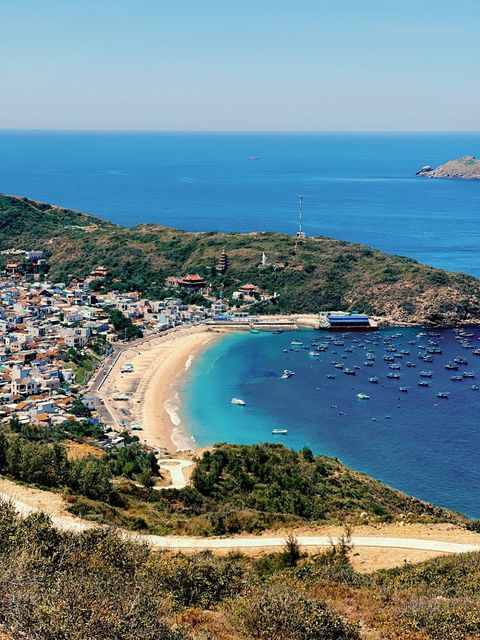 A fishing village on a curved bay, temples on the hill and blue boats on turquoise water, seen from a hillside path — shot on A CFX