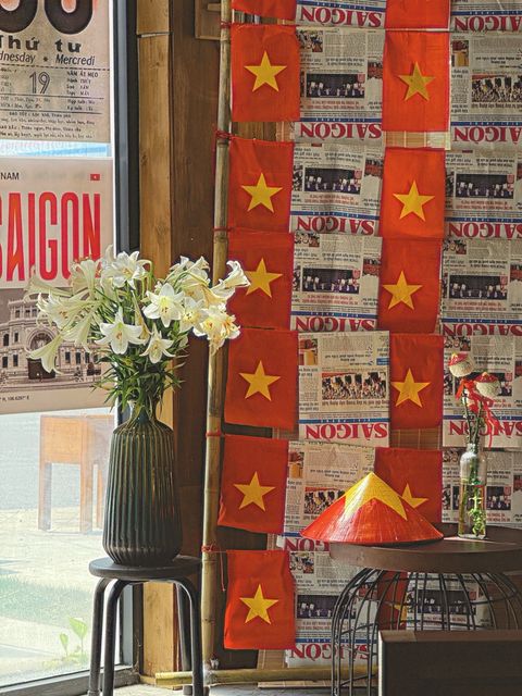 A café wall of red flags and old newspaper pages, white lilies in a vase by the window — shot on ELEV 02