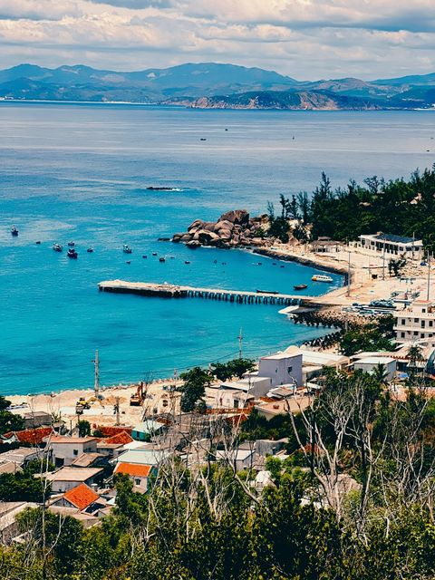 Seaside village with a long pier reaching into turquoise water, mountains on the horizon — shot on ELEV MAPLE